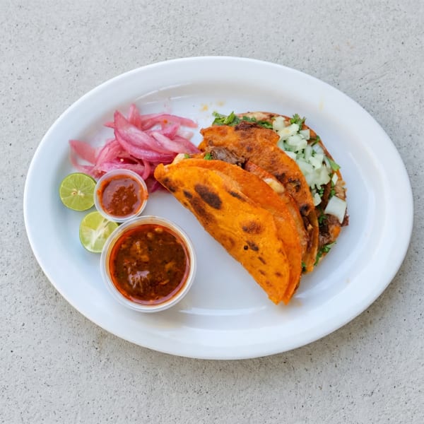 Three birria tacos with red onions, lime wedges, and two small cups of salsa on a white plate.